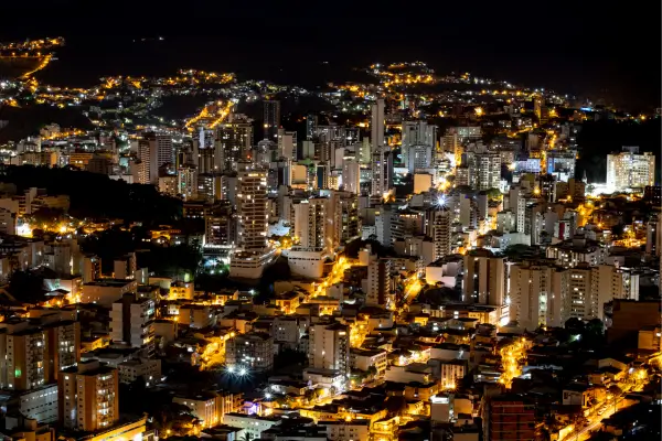 "Vista aérea noturna de Juiz de Fora, Minas Gerais, com prédios iluminados e ruas destacadas pelas luzes da cidade."