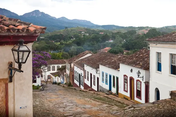 “Rua de paralelepípedos com casas coloniais preservadas em vilarejo histórico de Minas Gerais, com montanhas ao fundo.”